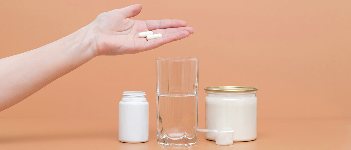 Eine Hand hält zwei weiße Kapseln über ein Glas Wasser. Links neben dem Glas steht die Umverpackung der Kapseln, rechts neben dem Glas ein Behälter mit weißem Pulver.