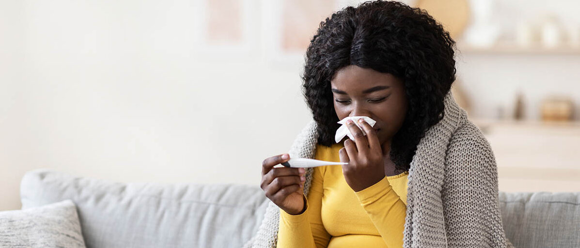 Eine schwangere Frau sitzt in eine Decke gewickelt auf dem Sofa, putzt sich die Nase und prüft ein Fieberthermometer.