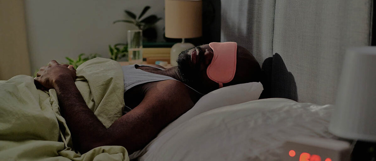 Long Covid Behandlung jetzt mit vier Medikamenten auf Kassenkosten Ein Mann liegt mit Schlafbrille im dunklen Zimmer im Bett.