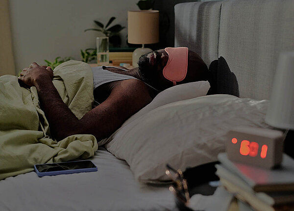 Long Covid Behandlung jetzt mit vier Medikamenten auf Kassenkosten Ein Mann liegt mit Schlafbrille im dunklen Zimmer im Bett.