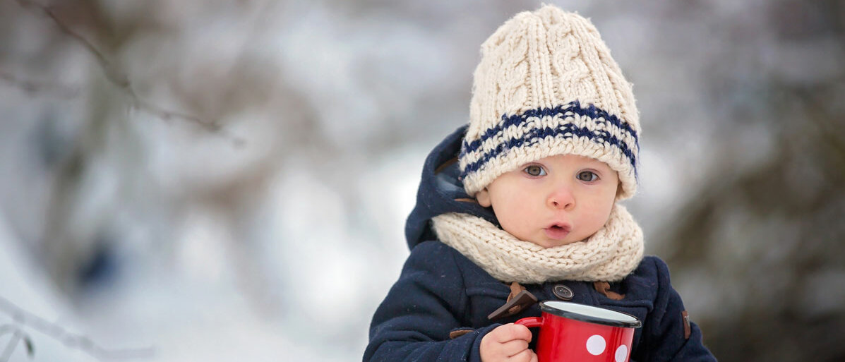 Kleines Kind steht dick eingepackt im Schnee und hat eine Tasse in der Hand