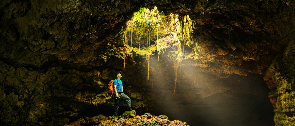 Ein Mann in Trekking-Kleidung steht in einer Höhle und schaut nach oben zu einer Öffnung, die er nicht erreichen kann.