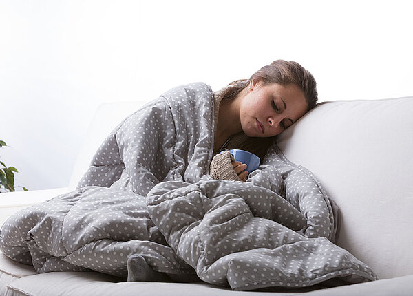 Eine Frau sitzt mit Decke und heißem Getränk auf dem Sofa und wirkt erschöpft.