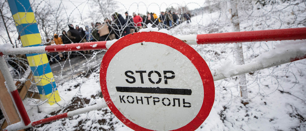 An einem Schlagbaum hängt ein Stopschild, in kyrillischer Schrift steht darauf "Kontrolle". Ein Pfahl links ist in den ukrainischen Farben blau und gelb gestrichen. Hinter einem Stacheldrahtzaun sieht man eine Menschenmenge Schlange stehen.