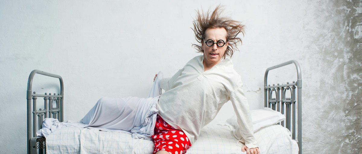 Ein Mann mit verwuschelten Haaren und runder Brille schwingt sich voller Elan aus dem Bett. Er trägt eine rote Boxershorts mit weißen Punkten, ein helles Shirt und schlüpft in blaue Crocs.