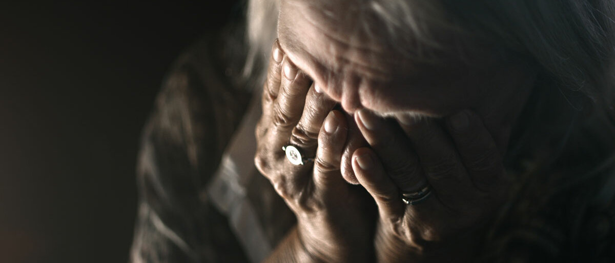 Eine sehr alte Dame sitzt im Dunklen und verbirgt ihr Gesicht in ihren Händen.