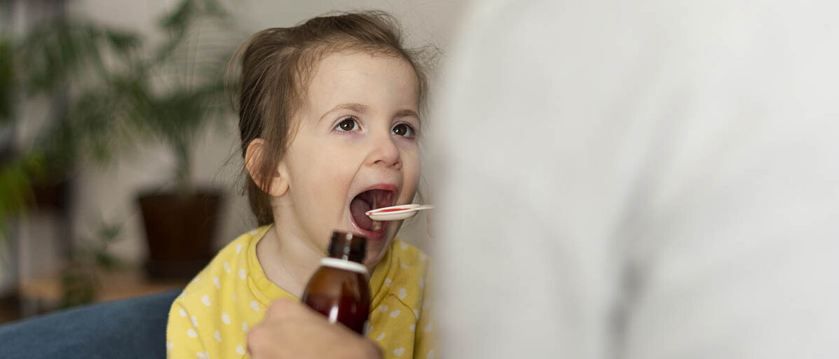 Dosierung bei Kindern: Dosierungsempfehlungen bei Off-Label-Therapien Ein Mädchen im Kindergartenalter hat den Mund weit aufgesperrt, um eine rosa Flüssigkeit von einem Dosierlöffel zu trinken.