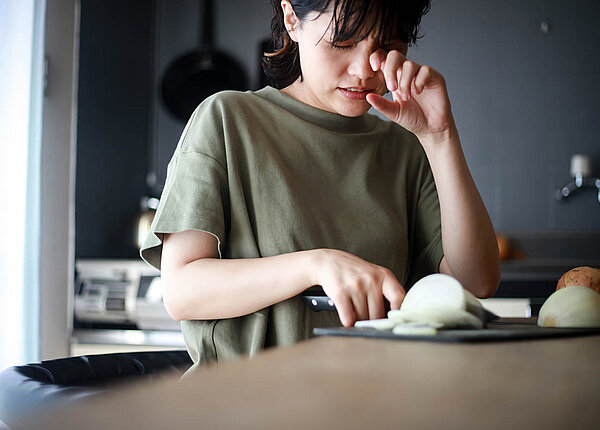 Eine junge Frau schneidet Zwiebeln. Sie kneift die Augen zusammen und wischt mit dem Fingerrücken eine Träne weg.