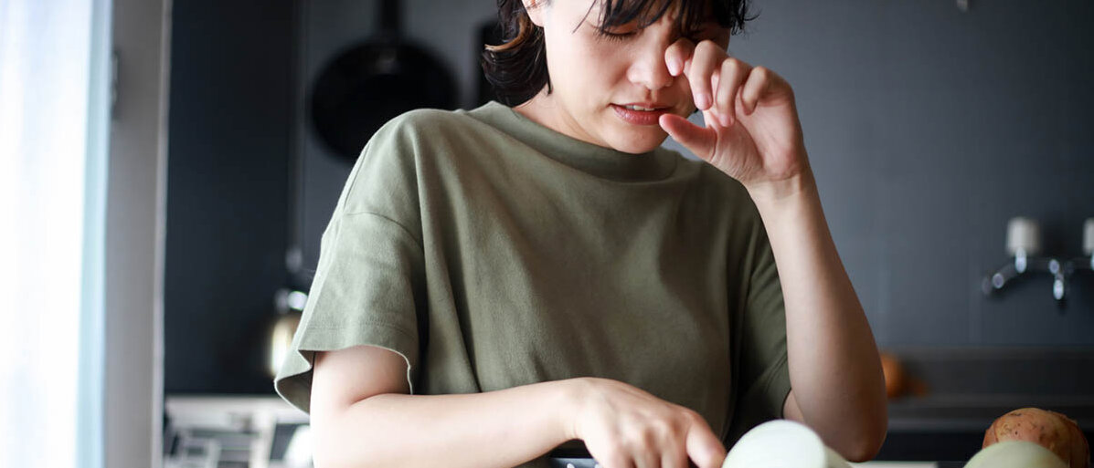 Tricks zum Zwiebelschneiden ohne Tränen Eine junge Frau schneidet Zwiebeln. Sie kneift die Augen zusammen und wischt mit dem Fingerrücken eine Träne weg.