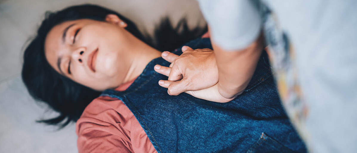 Erste Hilfe Maßnahmen: Notruf, Herzdruckmassage und wie man richtig hilft Eine Frau liegt regungslos und mit geschlossenen Augen auf dem Boden. Eine andere Frau gibt ihr mit verschränkten Händen eine Herzdruckmassage.