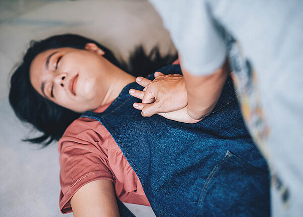 Eine Frau liegt regungslos und mit geschlossenen Augen auf dem Boden. Eine andere Frau gibt ihr mit verschränkten Händen eine Herzdruckmassage.