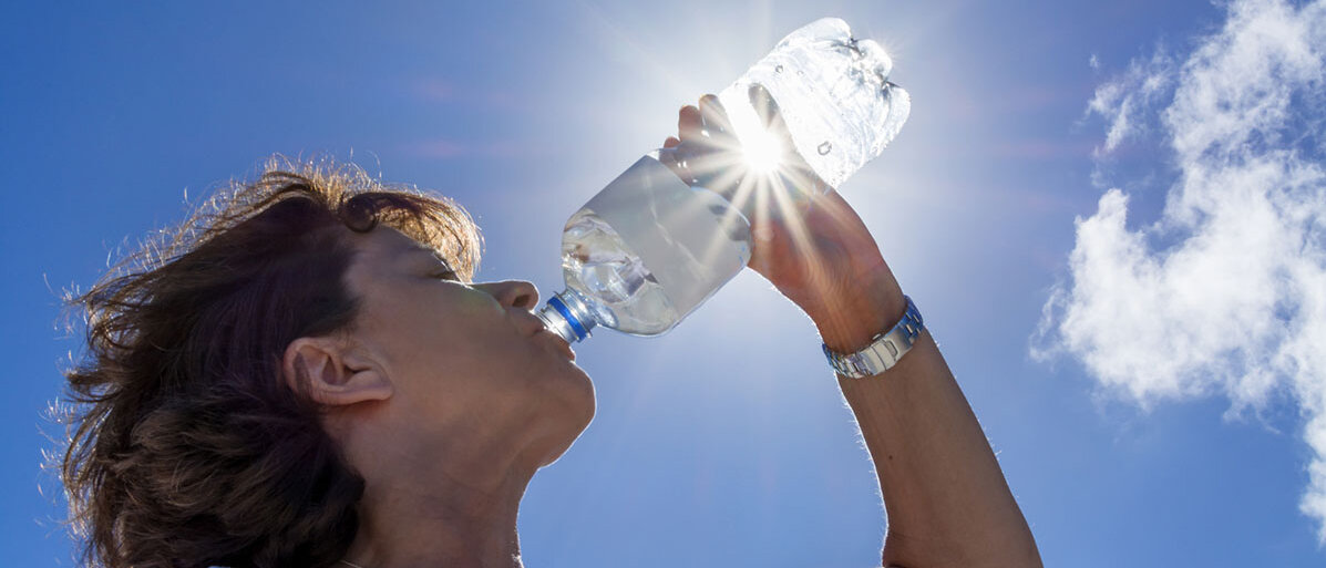 Ausreichend trinken bei Hitze Frau trinkt in der Sonne eine Flasche Wasser bei Sonnenstrahlen
