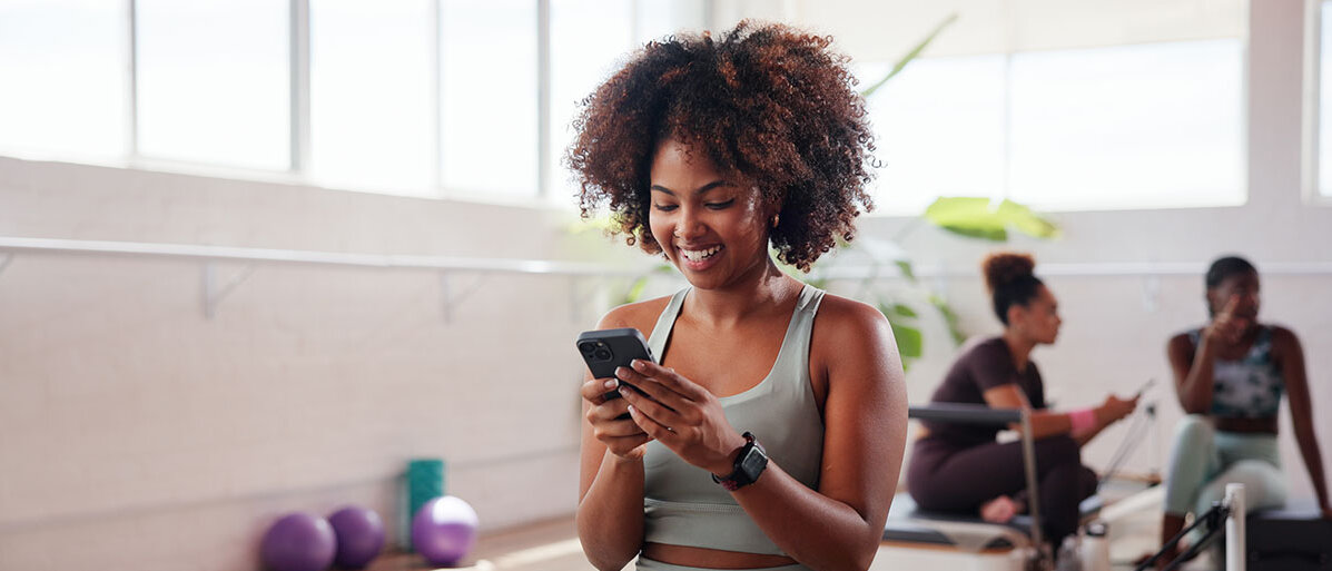 Glückliche afrodeutsche Frau mit Smartphone in der Hand im Fitnessstudio 
