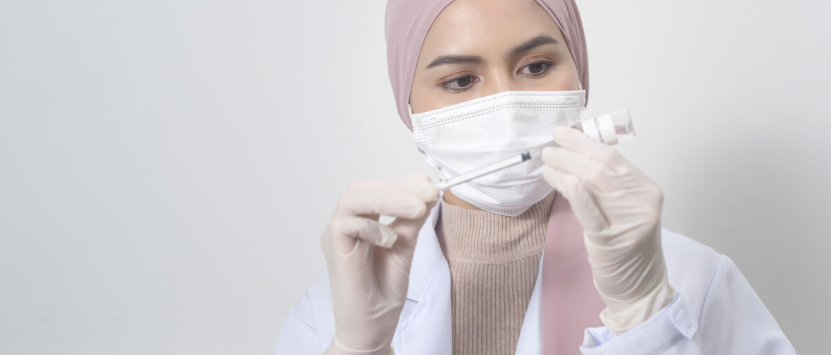 Eine junge Frau in Kittel mit Mundschutz und Handschuhen zieht eine Tuberkulinspritze mit der Flüssigkeit aus einem kleinen Glavial auf.