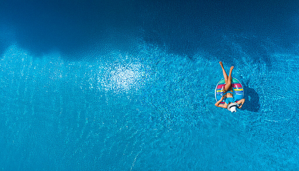 Ein Swimmingpool. Eine Frau liegt in einem großen Schwimmreif im Pool. Ihre Körpermitte hängt durch das Innere des Schwimmreifs hindurch im Wasser. 