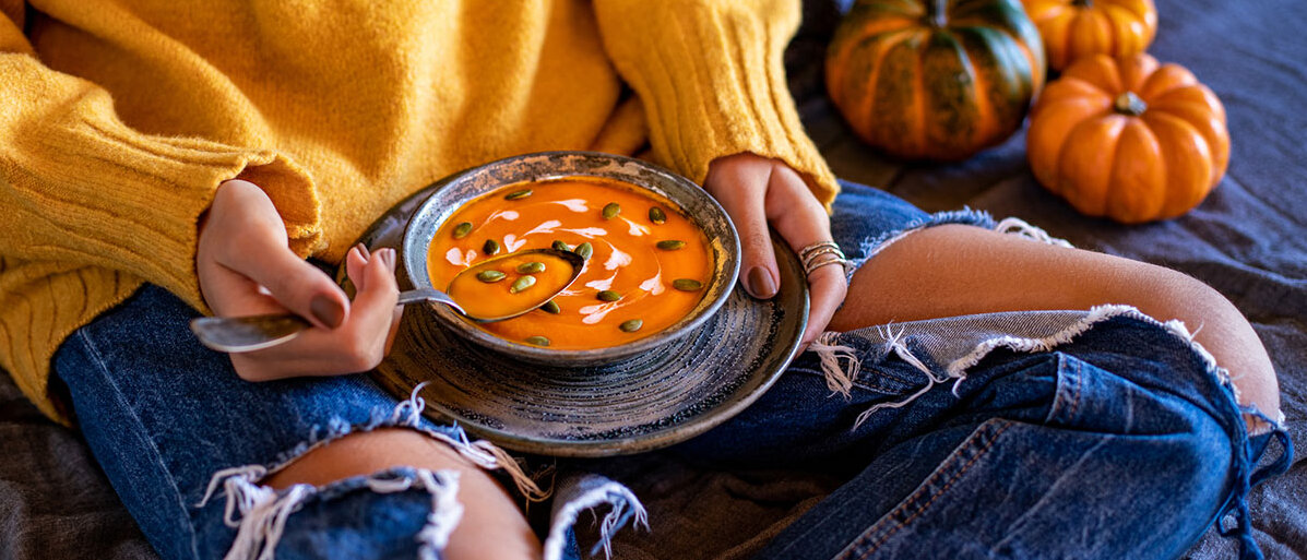 Frau in orangefarbenem Pulli auf dem Sofa mit einer Schüssel Kürbissuppe auf dem Schoß – neben ihr Zierkürbisse