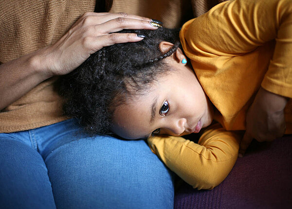 Ein kleines Mädchen liegt mit dem Kopf auf dem Schoß seiner Mutter, schaut erschöpft und hält eine Hand an den Bauch.