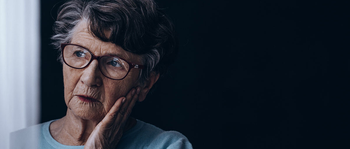 Eine ältere Dame steht in einem dunklen Raum, schaut aus dem Fenster und stützt verzweifelt das Gesicht in die Hand.