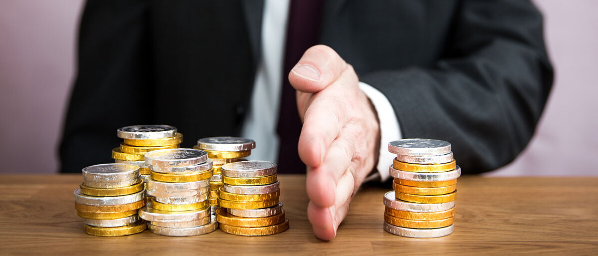 Vor einem Mann im Anzug stehen auf einem Tisch mehrere Stapel mit Münzen. Der Mann teilt mit der Hand einen der Stapel ab und schiebt ihn zur Seite.