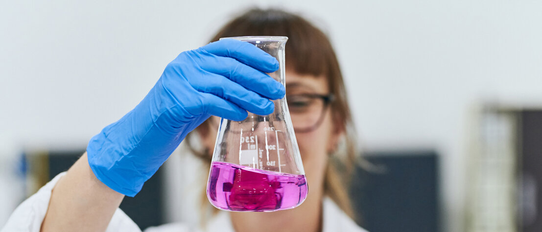 Eine Frau im Laborkittel und mit Handschuhen hält einen Erlenmeyerkolben hoch, darin ist eine violette Lösung.