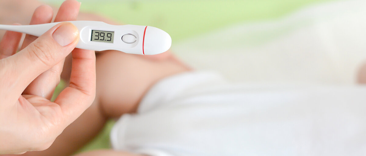 Ein Fieberthermometer zeigt 39,9°C, im Hintergrund ist ein liegendes Baby
