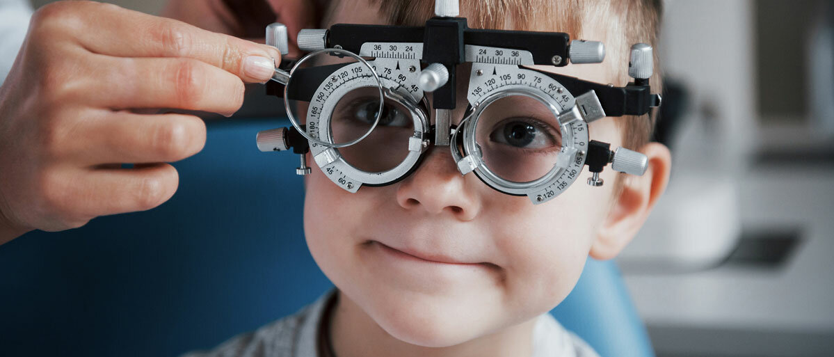 Ein Junge im Vorschulalter bekommt eine Brille angepasst. Dazu trägt er einen Optometrie-Rahmen, ein brillenartiges Gestell, in das die Optikerin oder Augenärztin verschiedene Gläser einlegt.