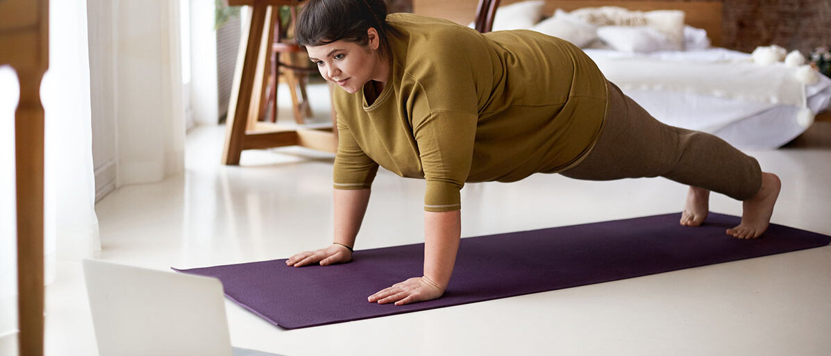 Eine Frau macht eine Übung auf einer Yogamatte. Dabei schaut sie in ihren Laptop, daneben liegt ihr Smartphone.