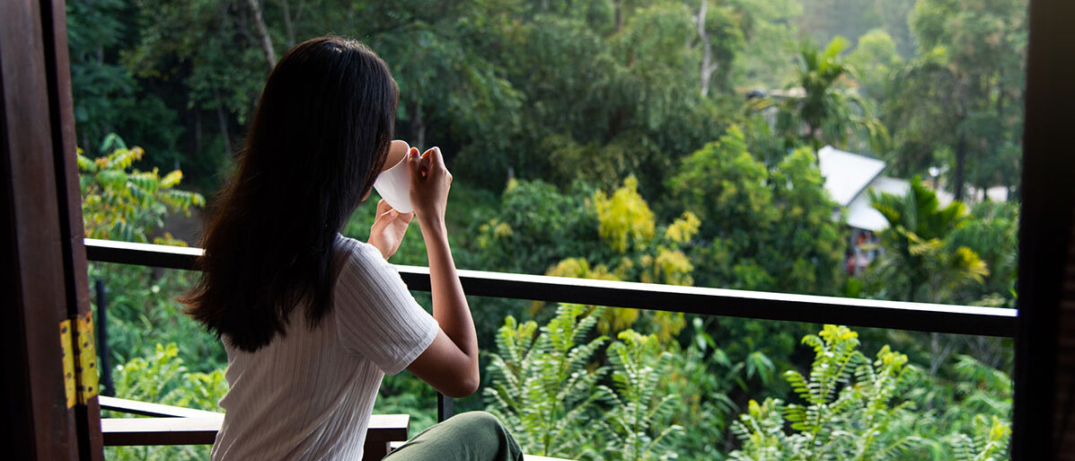 Eine dunkelhaarige Frau sitzt auf einem Balkon, blickt auf einen dichten Wald und trinkt aus einer Tasse.