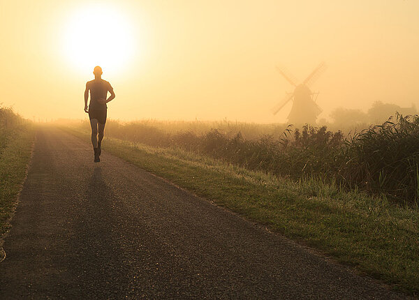 Sport bei Hitze Mann, der in der nebligen, niederländischen Landschaft in der Nähe einer Windmühle läuft.