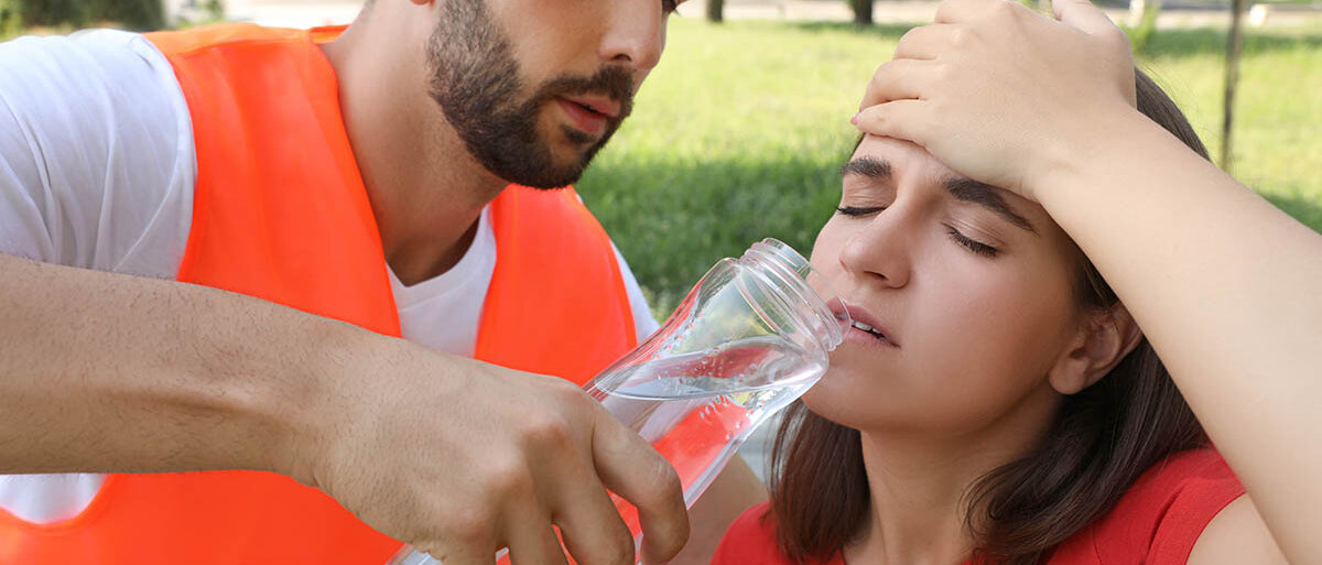 Erste Hilfe bei Hitzschlag und Sonnenstich: Symptome erkennen und Maßnahmen ergreifen Ein Sanitäter reicht einer Frau eine Flasche Wasser. Sie sitzt auf dem Boden, schaut gequält und hält sich die Hand an den Kopf.