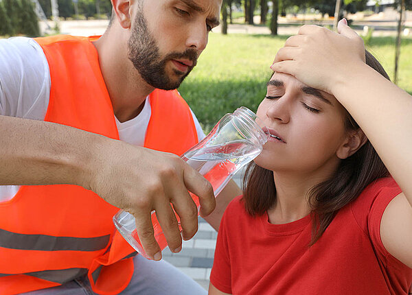 Erste Hilfe bei Hitzschlag und Sonnenstich: Symptome erkennen und Maßnahmen ergreifen Ein Sanitäter reicht einer Frau eine Flasche Wasser. Sie sitzt auf dem Boden, schaut gequält und hält sich die Hand an den Kopf.