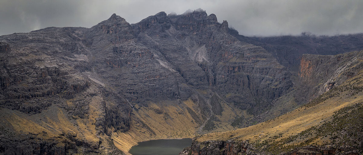 Blick ins Gorgeous Valley des Mount Kenya in Kenia: grün bewachsene Hänge, steinige Steilhänge, ein See, Wolken.