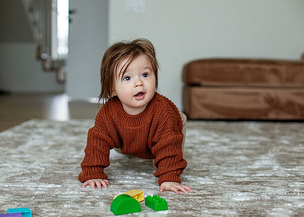 Schadstoffe im Hausstaub ein Risiko für Babys und Kleinkinder Kleines Mädchen mit braunem Pullover krabbelt auf dem Teppich.