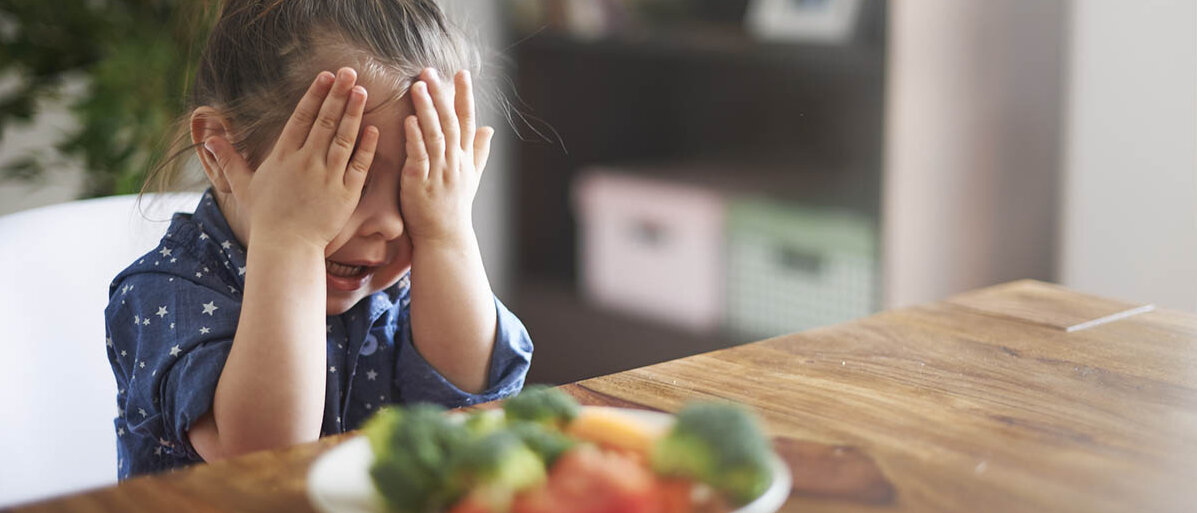 Ein kleines Mädchen sitzt am Tisch und hat einen Teller mit Gemüse vor sich. Das Mädchen hält sich die Augen zu.