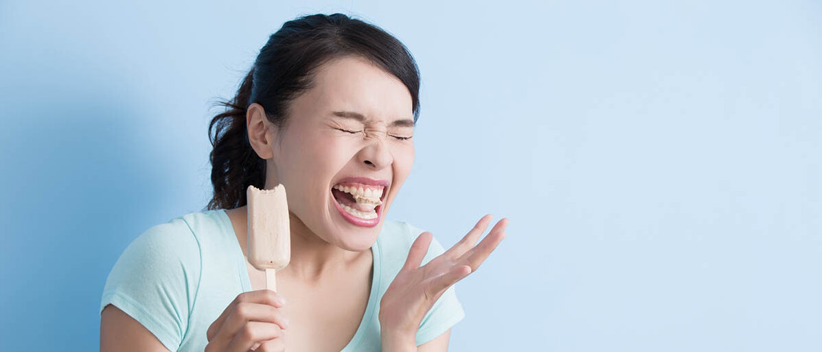 Zahncreme für empfindliche Zähne und andere Tipps für freiliegende Zahnhälse, um den Zahnschmelz zu schützen. Eine Frau hat in ein Eis am Stiel gebissen und verzieht das Gesicht.