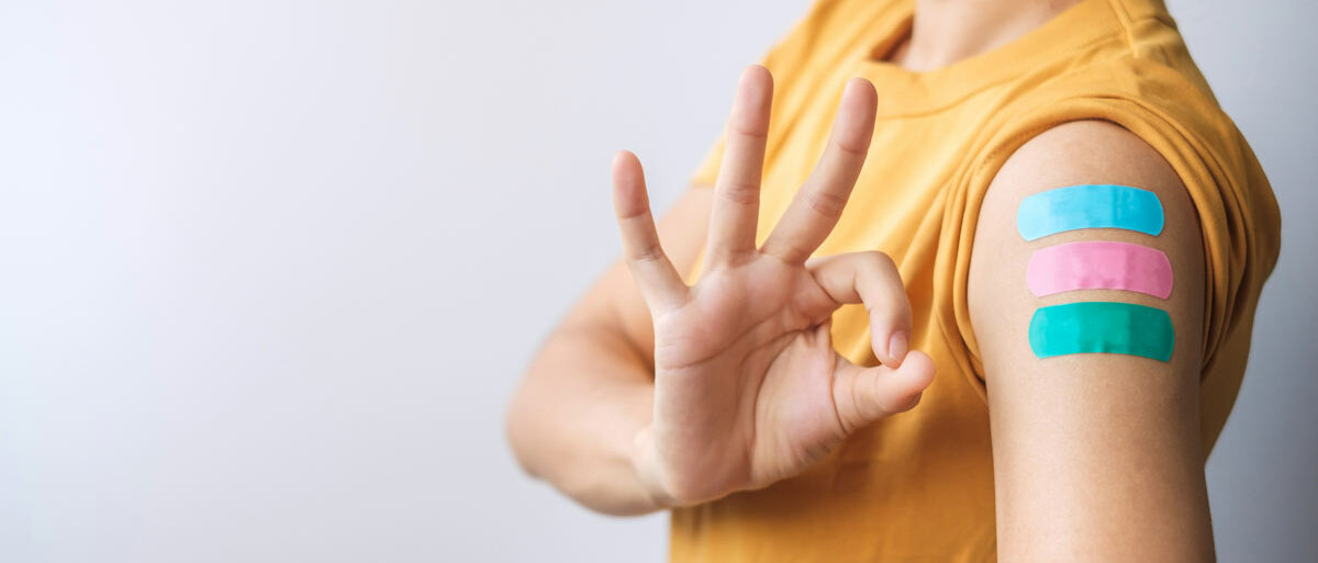 Eine Person mit chirurgischer Gesichtsmaske hat den Ärmel ihres Shirts hochgezogen. Auf dem Oberarm sind drei Pflaster in jeweils unterschiedlicher Farbe zu sehen. Die Person bildet mit Daumen und Zeigefinger der anderen Hand einen Kreis als "OK"-Geste.