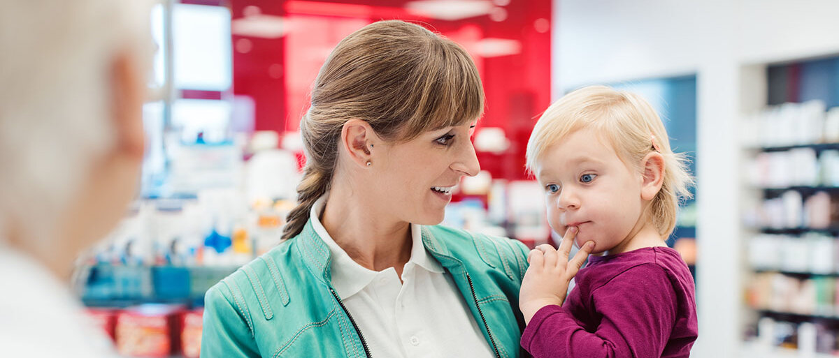 Eine Frau hält ein etwa zweijähriges Kind auf dem Arm. Sie steht in einer Apotheke am Handverkaufstresen eine*r Apothekenmitarbeiter*in gegenüber.