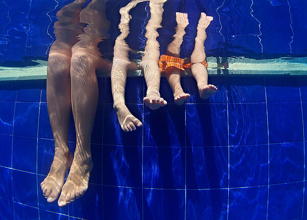Wir blicken unter Wasser auf drei Beinpaare: Eine Mutter und ihre zwei Kinder lassen die Beine im Wasser baumeln.