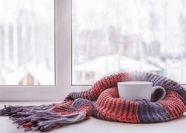 Erkältung: Husten und Halsschmerz Auf einer Fensterbank steht eine dampfende Tasse, um die ein Strickschal geschlungen ist. Draußen liegt Schnee.