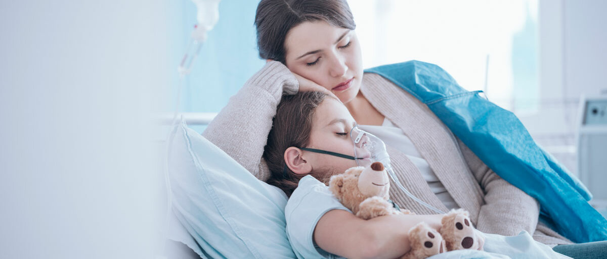 Ein Kind liegt mit Sauerstoffmaske in einem Krankenhausbett und kuschelt sich an seinen Teddy und seine Mutter.