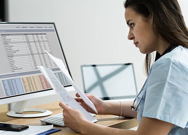 Eine Frau im Kasack und mit umgehängtem Stethoskop sitzt am Computer. Der Bildschirm zeigt Tabellen mit Kalkulationen. Außerdem hält sie Papiere in der Hand.