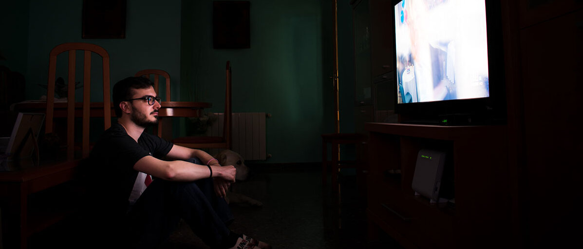 Mann sitzt vor dem Fernsehen, der Raum ist dunkel, man sieht nur das Licht des Fernsehers