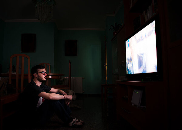 Mann sitzt vor dem Fernsehen, der Raum ist dunkel, man sieht nur das Licht des Fernsehers