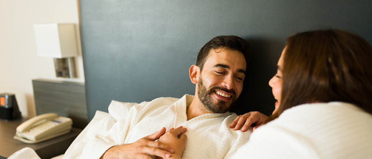 Ein Mann und eine Frau liegen im Bademantel im Bett. Die Frau hat ihre Hand auf die Brust des Mannes gelegt, er hält ihre Hand fest und beide lächeln sich an.