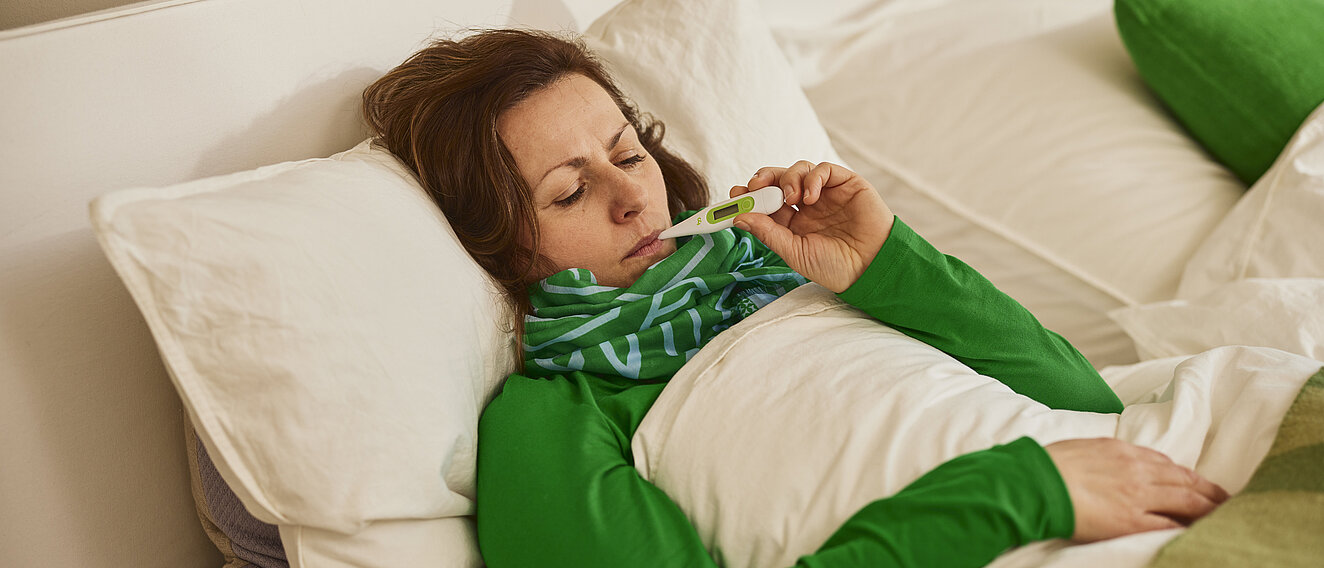 Eine Frau liegt im Bett und misst ihr Fieber mit einem Fieberthermometer im Mund