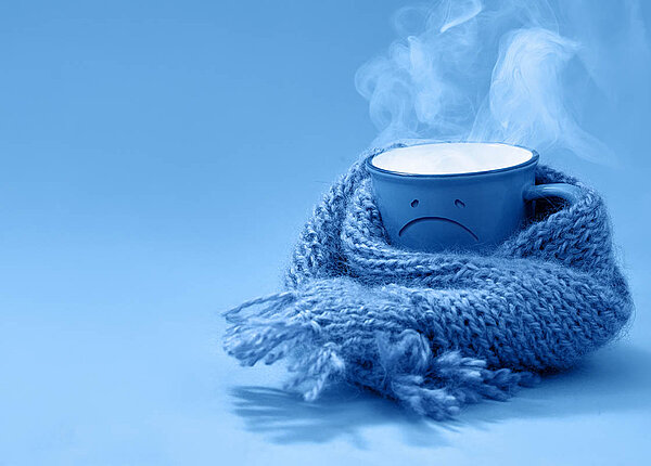 Ursache für Schüttelfrost: Warum wir bei Fieber frieren eine Tasse mit dampfendem Inhalt hat ein motziges Gesicht und einen Schal umgewickelt