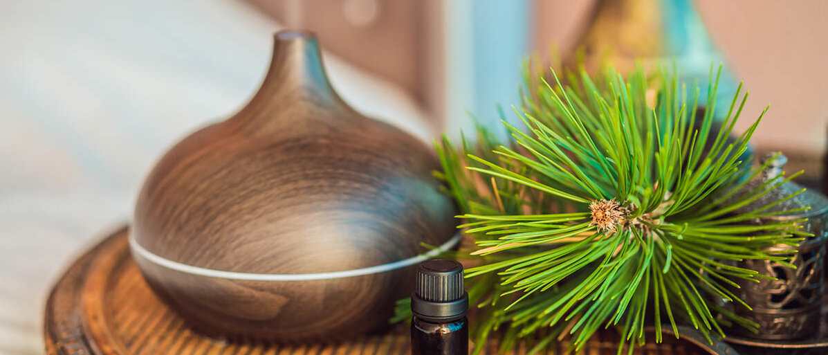 Aroma-Diffuser in Zwiebelform und Holzoptik auf einem Beistelltisch, daneben ein Zweig eines Nadelbaums und ein Braunglasfläschchen mit ätherischem Öl.