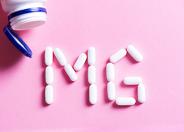 Flatlay: Aus einer Tablettendose sind weiße Tabletten herausgefallen und bilden die Buchstaben „Mg“.