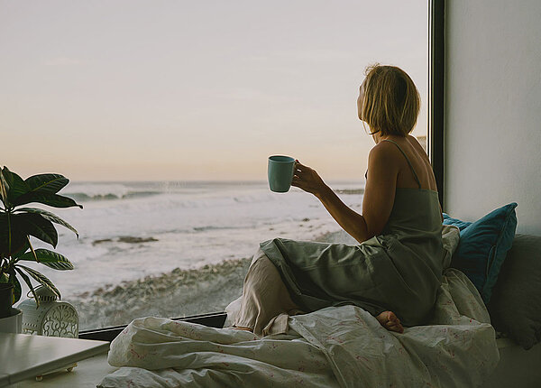 Frau sitzt mit einer Tasse entspannt auf ihrem Bett am Fenster. Sie schaut aus einem Panoramafenster aufs Meer