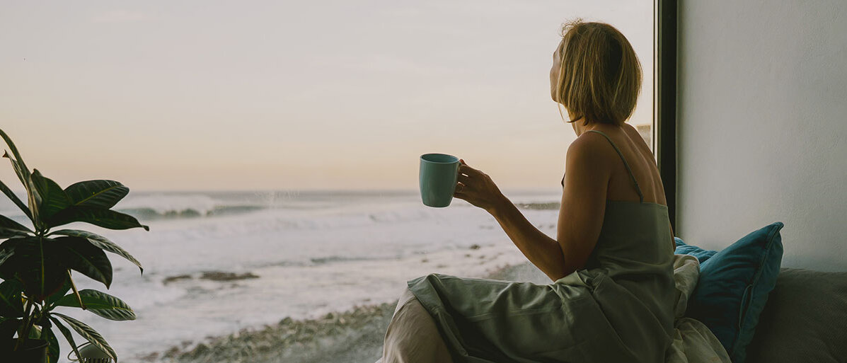 Durch Kurzurlaub Stress abbauen und Erholung starten Frau sitzt mit einer Tasse entspannt auf ihrem Bett am Fenster. Sie schaut aus einem Panoramafenster aufs Meer